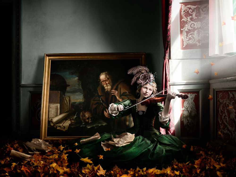 En kvinna i grön klänning och en huvudbonad med fjädrar sitter på goolvet och spelar fiol. PÅ marken finns höstlöv och bakom kvinnan står en målning. 