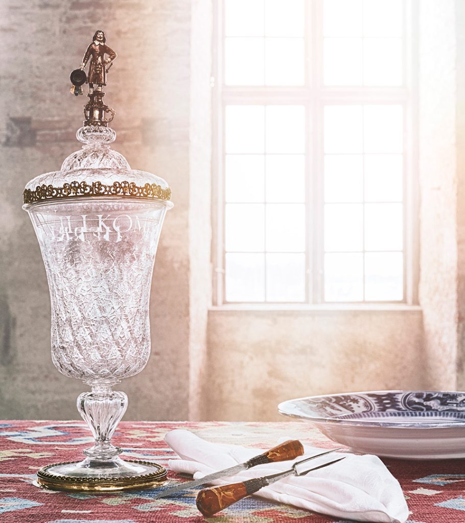  A glass trophy on a table next to a plate, cutlery and napkin. On top of the cup is a sculpture of a man in 17th century clothing.