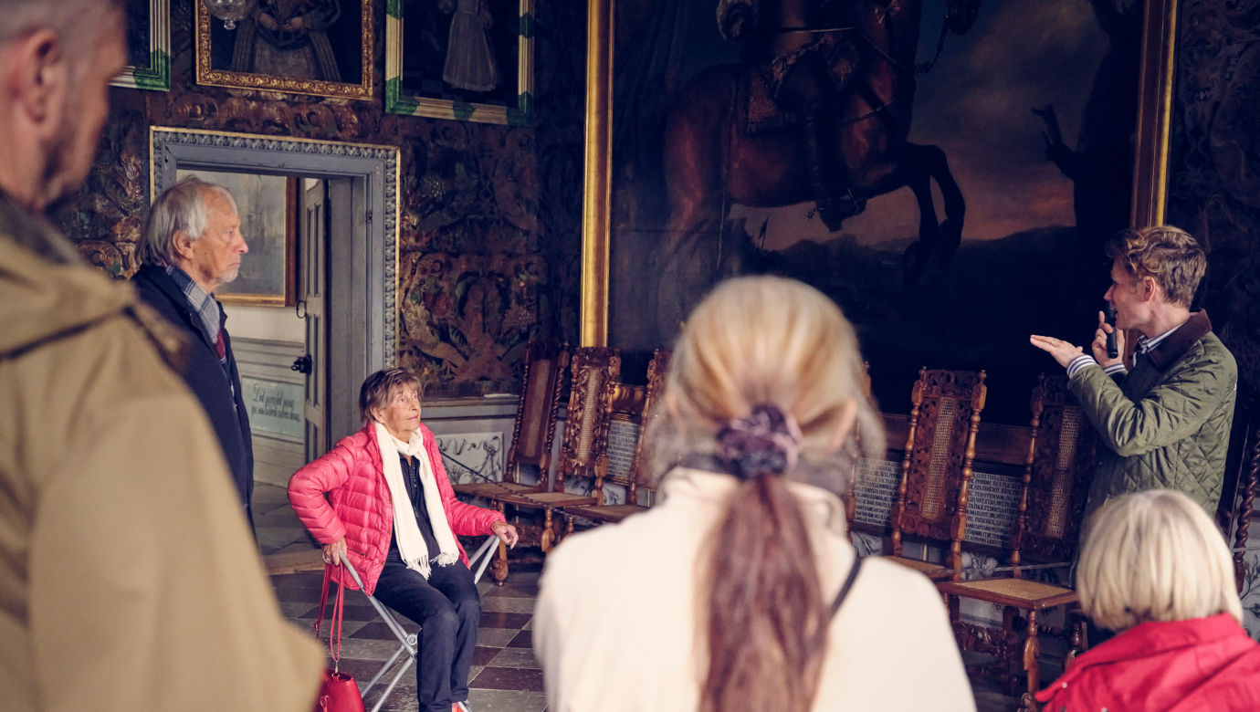 Kvinna som sitter på en bärbar stol och lyssnar på en guide i ett av slottets rum.