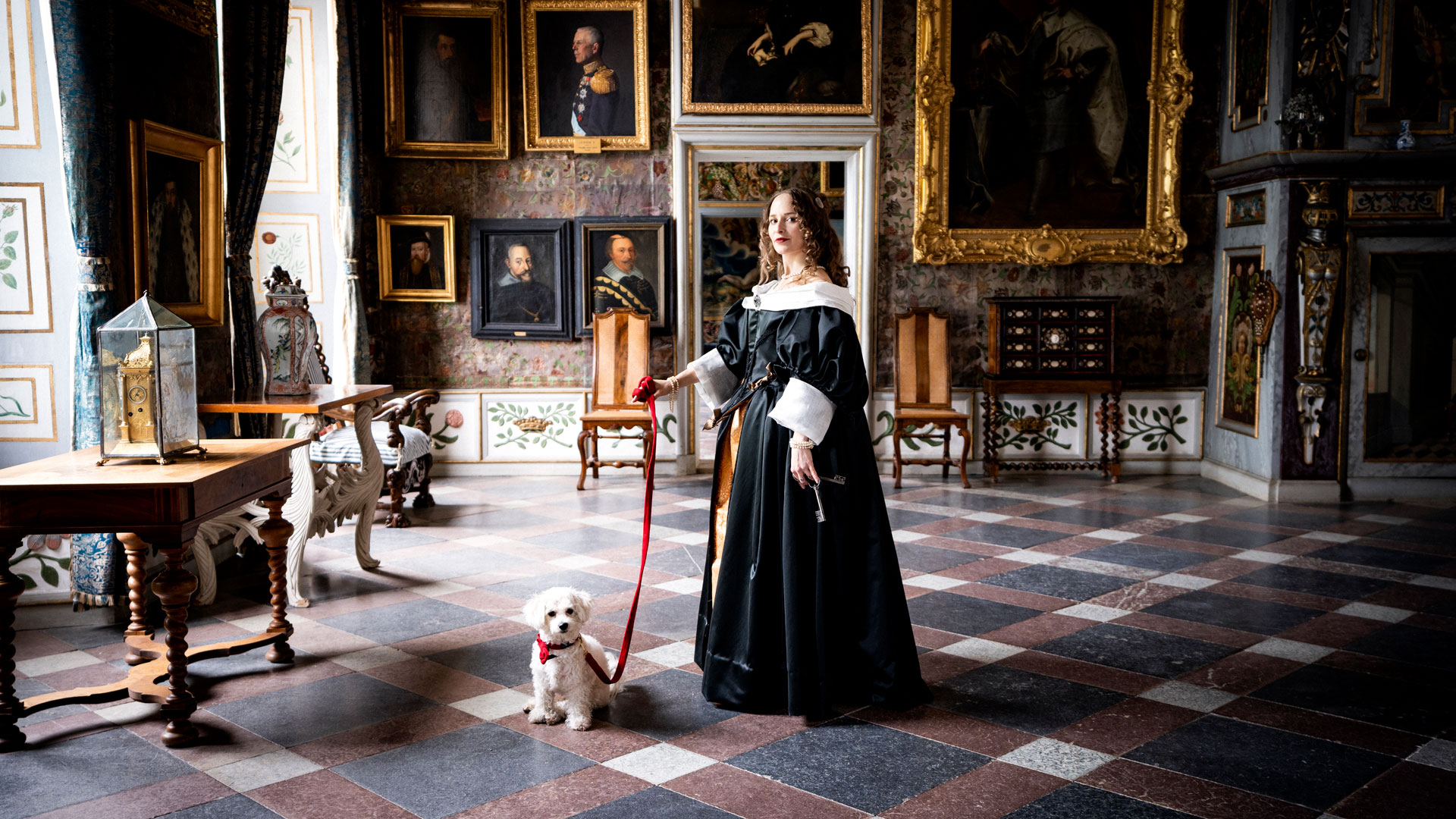 Woman dressed as a castle lady stands in the King's Hall with a small white dog on a red leash.