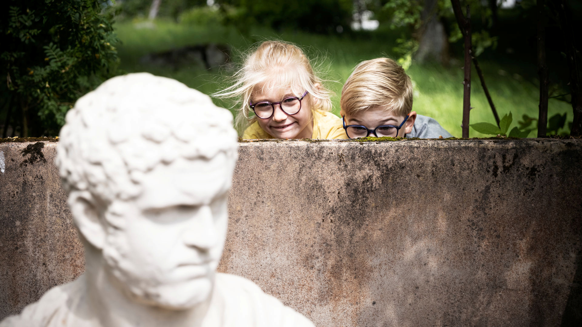 Two children peek out from behind a wall to look at a white statue.