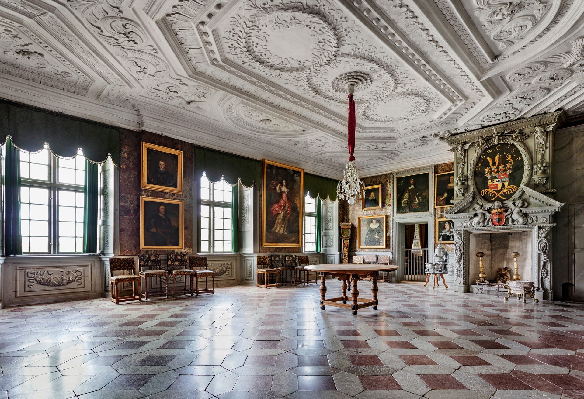 A very large room with patterned flagstone floor, lavishly decorated fireplace and several paintings.