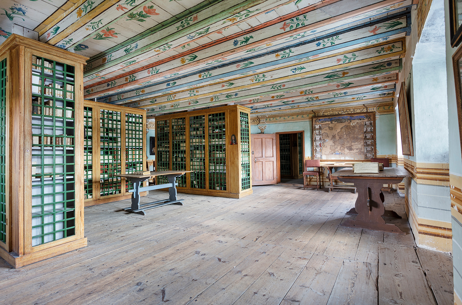 A room with bookshelves. The bookshelves have green grill doors. The ceiling is painted with flowers.