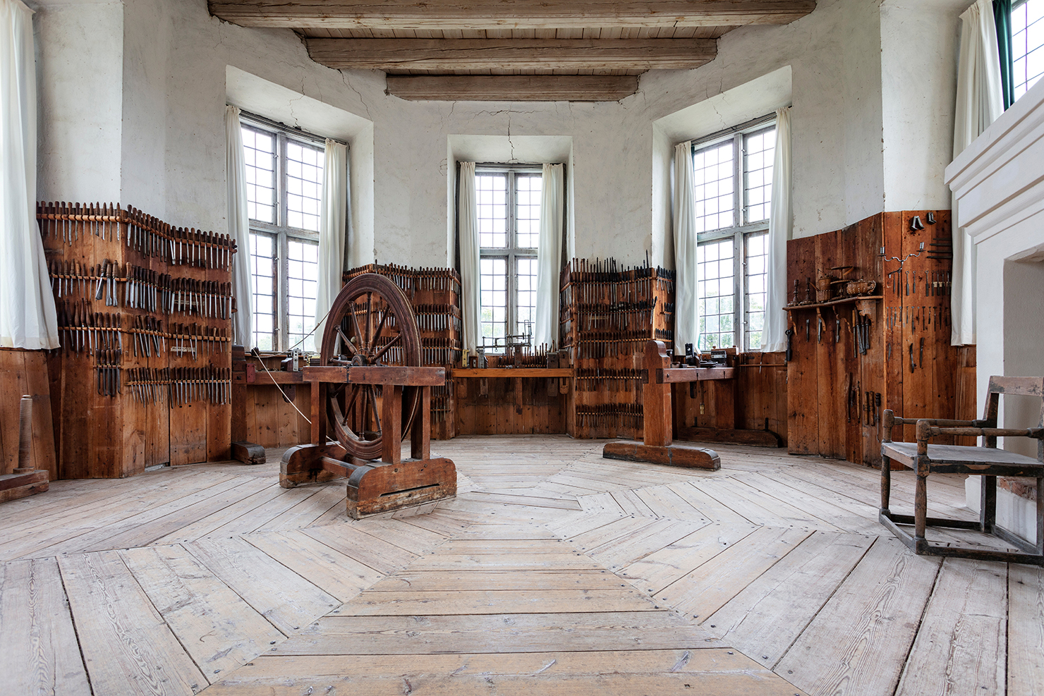 A partially wood-panelled room with a large wooden wheel in the middle. A number of wood-working tools are hanging on the walls.