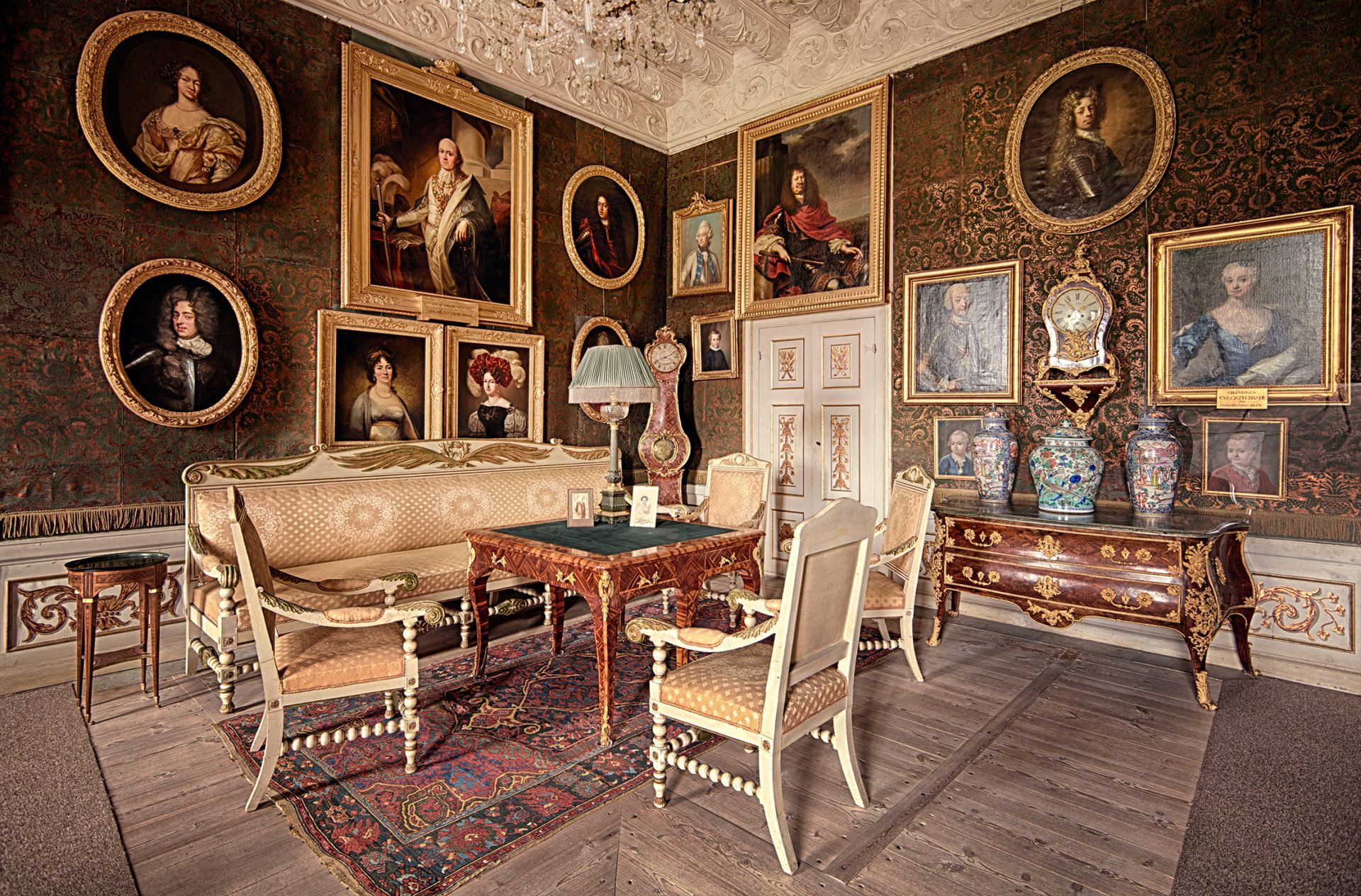 Ett tätt möblerat rum med möbler i vitt och guld. På väggarna hänger en mängd porträtt i guldramar.