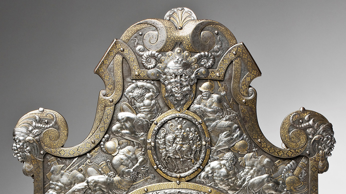 Detalj av en sköld av delvis förgyllt stål. Skölden är rikt dekorerad med bundna krigsfångar och ett leende, hornförsett djävulshuvud..