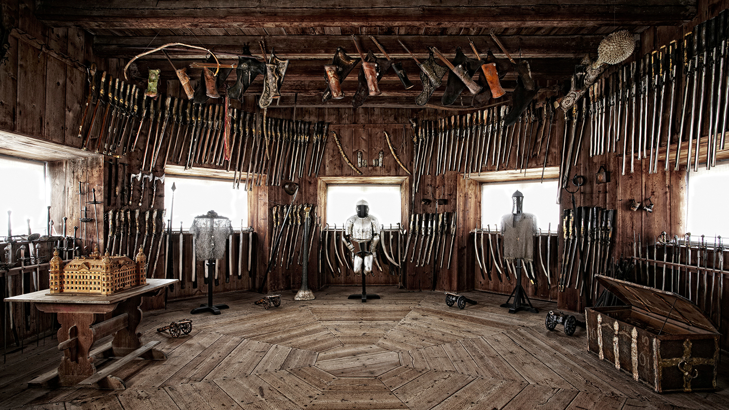 An octagonal, wood-panelled room with low windows. The room is filled with weapons and suits of armour.