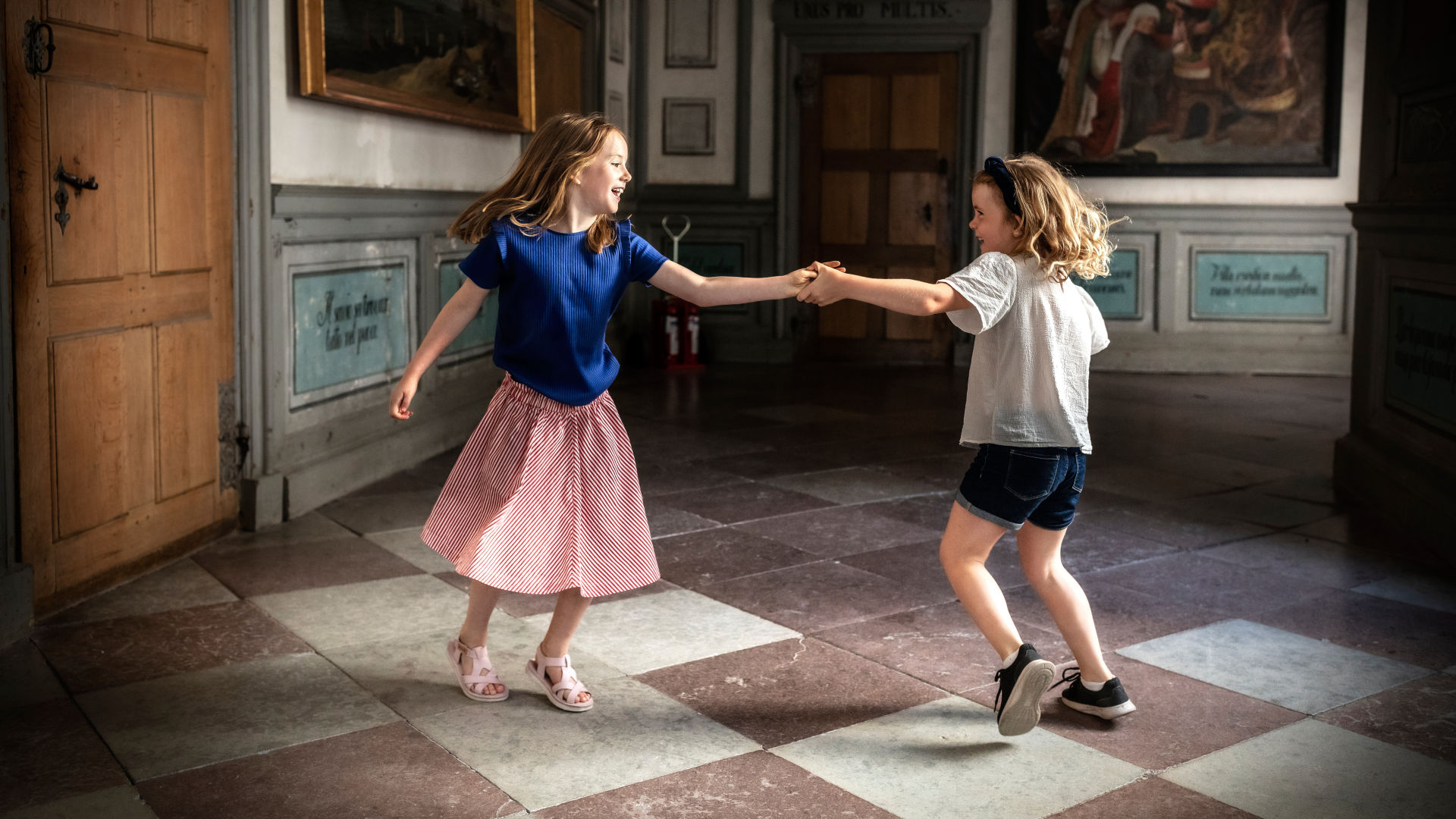 Två barn håller varandra i handen och dansar i slottets korridor