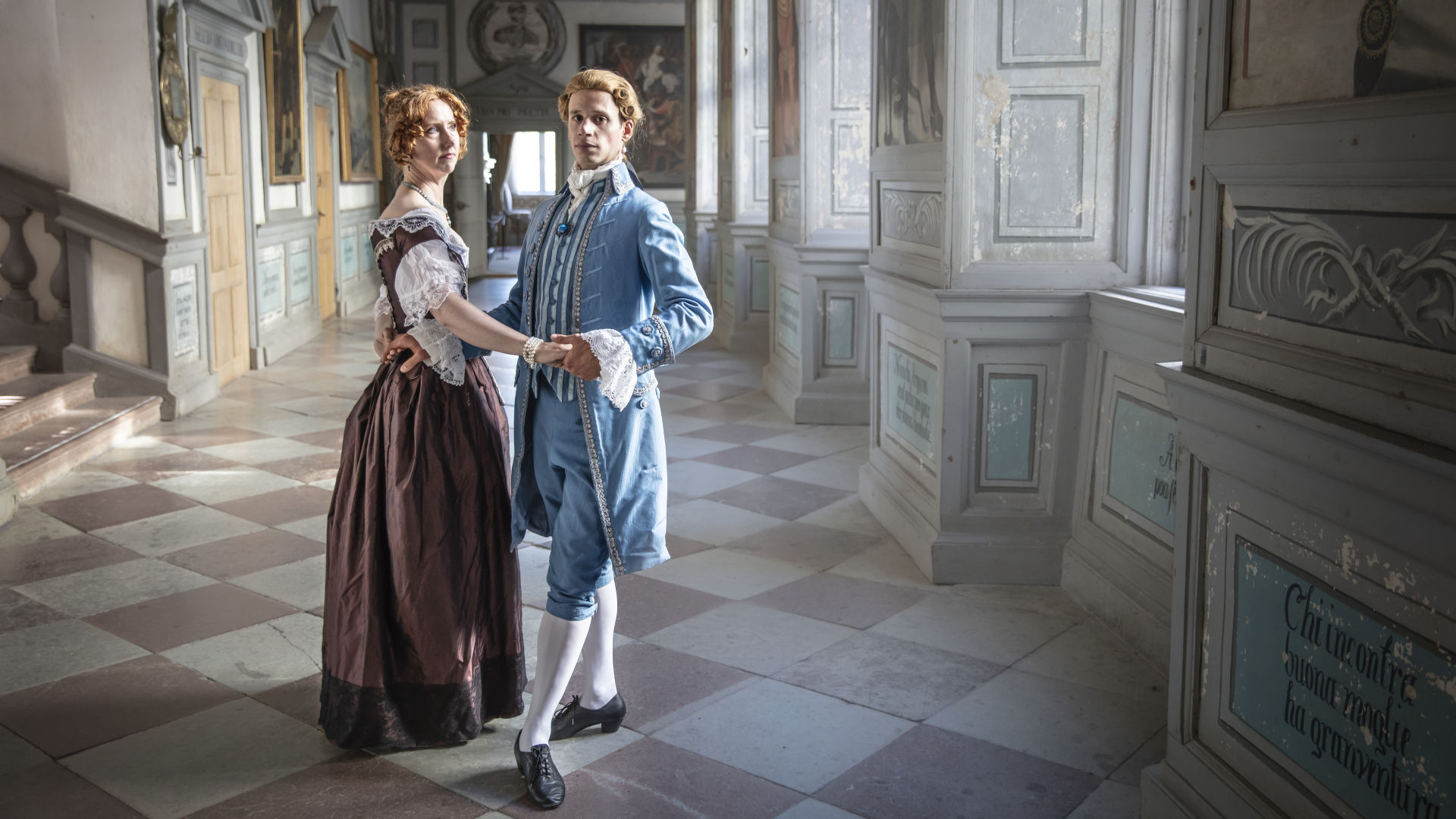 Två personer i historisk dräkt står i dansposition i slottets korridor.