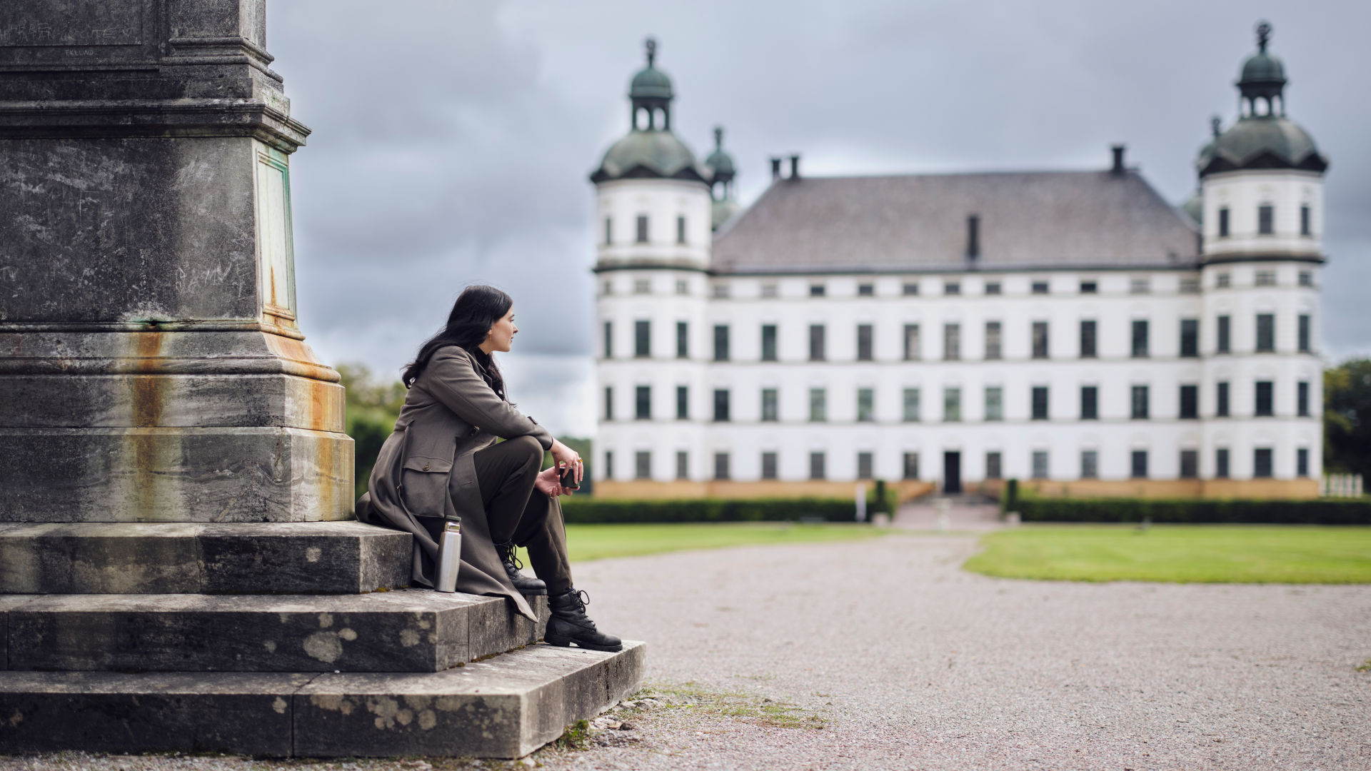 En person som sitter på en trapp i vänster i bilden och tittar ut på slottsparken och slottet.