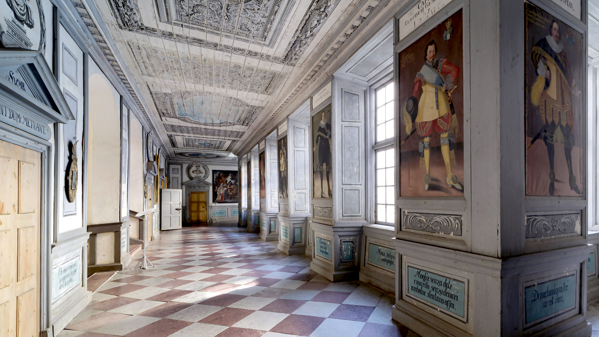 En lång korridor med schackrutigt stengolv i ljusgrått och tegelrött. Längs korridorens ena sida fönster, längs andra dörrar. På väggarna mellan fönstren finns stora 1600-talmålningar som föreställer män i vackra kläder.