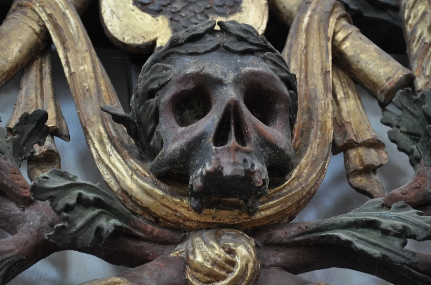 En närbild av en skulpterad dödskalle i trä, målad i mörka toner med spår av rött och svart. Skallen är omgiven av guldfärgade band och dekorativa bladornament i grönt och brunt. Figuren är en del av en barock utsmyckning och har ett dramatisk och symboliskt uttryck.